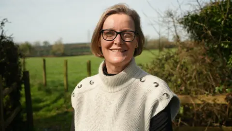 Tessa is looking at the camera with a slight smile. she has on black rimmed glasses and a sleeveless beige turtleneck sweater with brown buttons on the shoulders. Behind her is a countryside scene with hedgerow brambles on either side of a path marked by wooden posts and fencing arounda green field with solar panels out of a focus on the horizen