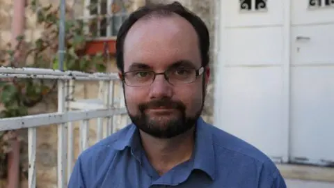 Symon Hill Photograph of Symon Hill, a white man with a short beard and glasses wearing a blue shirt