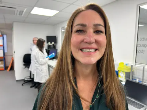 A long haired woman smiles at the camera with a laboratory behind.