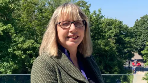 Nicola Day has shoulder length blonde hair. She is standing outside on a balcony amongst leafy green trees and a blue sky. She is wearing a green jacket and blue shirt. She is looking directly at the camera and is wearing glasses.
