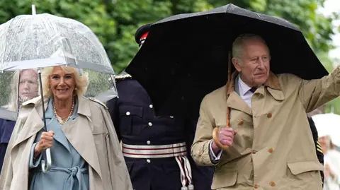 Queen Camilla in a blue dress with a beige trench coat holding an umbrella walking towards the camera, with King Charles III in a brown coat holding a black umbrella and waving.
