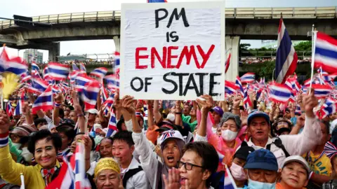 A woman standing in the centre of a crowd that fills the image, holding a placard that reads "PM is enemy of state" in black lettering, with enemy written in red. The entire crowd is filled with people waving the Thai flag. In the background is an overpass.