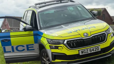 A yellow, blue and white liveried police car with a bullet hole in the windscreen sits on a grassy bank in a housing estate. The driver's door is lying open.