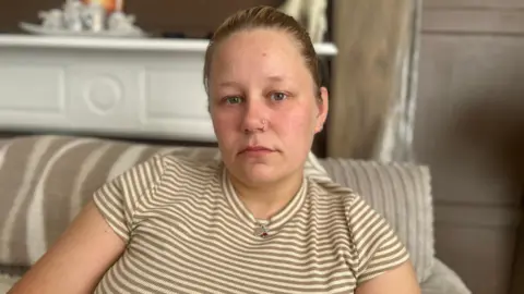 BBC A person sits on a striped sofa in a living room, facing the camera. The person is wearing a short-sleeved beige and white striped top, a necklace, and a nose stud. Tattoos are visible on both forearms. The background includes a white mantelpiece and wooden wall panels, with a shallow depth of field keeping the person in focus while the room behind is softly blurred.