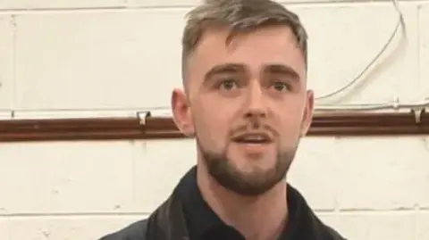 Conor Reddy Councillor Conor Reddy, a man with short, fair hair and a beard, speaks into a microphone. He is wearing a black jacket over a black shirt. 