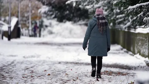 PA A woman wrapped up in a winter coat, scarf, hat and gloves walks along a snowy pavement