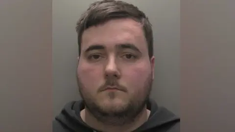 A police custody shot of a man with short brown hair and brown eyes. He has a thin stubbly beard and is wearing a black hooded sweatshirt.