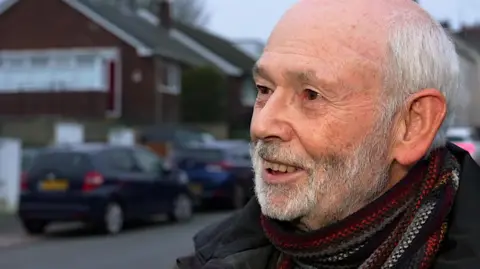 BBC David Radford, with grey hair and beard, wearing a black and red scarf.