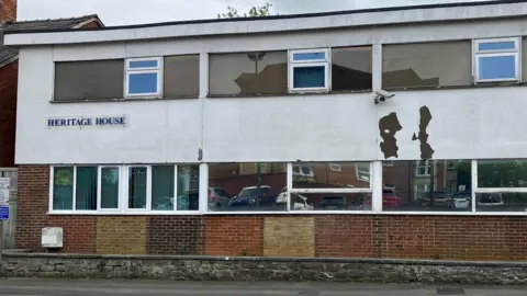 An ageing building with peeling paint and a security camera. It says 'heritage house' on it.