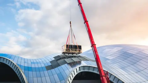 Ben Hines A large chiller unit is strapped to a red crane and lifted out of The Glasshouse where glass panels have been removed from the roof of the building. The orange, blue and grey sky is reflected on the curved glass roof of The Glasshouse. 