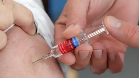 A man receiving a vaccination into his arm. He is wearing a white t-shirt while the medic is pressing down on the syringe to administer the vaccine.