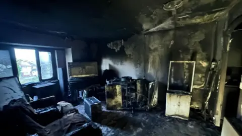 The aftermath of the fire in the flat. A living room is burnt-out. There are smoke-blackened walls and charred items of furniture.