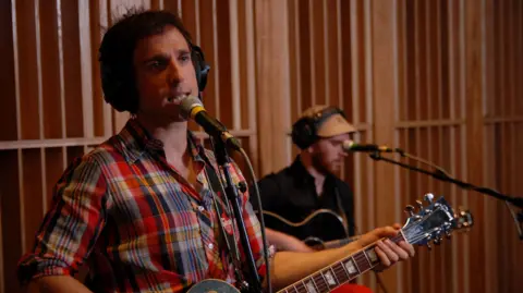 DAVID HOOPER/BBC Hoosiers vocalist Irwin Sparkes is wearing a checked shirt and black headphones and singing into a microphone on a stand while holding a guitar. Behind him, Alan Sharland is out of focus, wearing black headphones over a cap and a black shirt. He is also holding a guitar and has a microphone in front of him. Behind them is a wall made of vertical slats of wood.