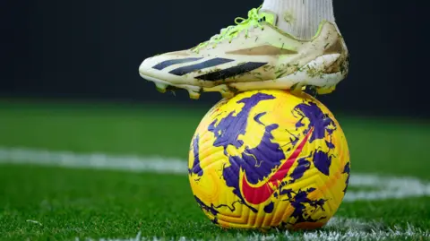 A football boot resting on a football on a football pitch.