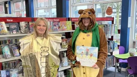 Warwickshire Library & Information Service The image shows two people standing inside a brightly lit library, surrounded by children’s books displayed on shelves.
In the foreground, one person is dressed in a full-body brown animal costume resembling a bear and is holding a children’s book with an illustrated cover. The other person is wearing a large gold, chocolate-bar-style costume with visible text including “GOLDEN TICKET” and smaller printed details.