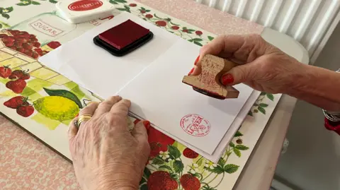 A pair of woman's hands with red nail varnish holding a stamp featuring Lover village above a white envelope.