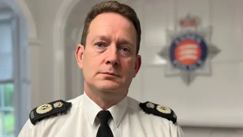 Stuart Woodward/BBC Chief Constable Ben-Julian Harrington, who has short light brown hair. He is wearing a white shirt with blue police lapels and a dark tie. He is sitting in a room with the Essex Police log blurred out on a white wall behind him.