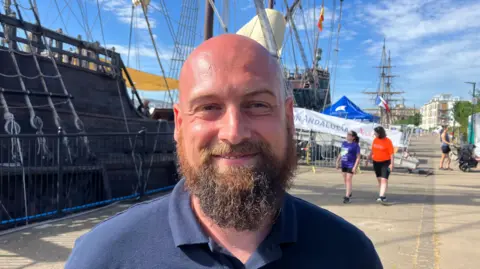 A smiling Lupo Bathke stands on a quay in front a replica Spanish galeon. He is smiling and wearing a blue polo shirt. He is bald, two women can be seen in the background