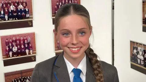 John Devine/BBC Hermione has long brown hair which is in a plait. She is wearing a white shirt, grey blazer and blue tie and is standing in front of a display which is showing several photographs.