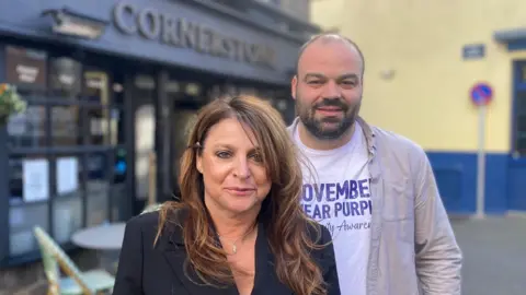 Jo is on the left of the picture. She is wearing a navy blue suit jacket and is standing in front of Marvin who is wearing a white t-shirt with a beige unbuttoned shirt on top.