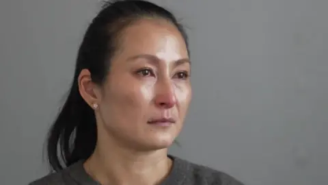 Head and shoulders of Tina Campbell, she has long dark hair tied in a ponytail and is visibly upset. She is sitting in front of a plain grey background and wearing a dark grey sweatshirt