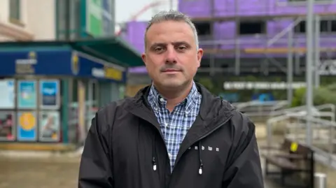 Carl Walker, a man with grey hair and grey stubble. He is wearing a shirt with blue and white checks, underneath a black hooded coat with a zip through the middle. He is standing outside a Coop supermarket in St Helier.