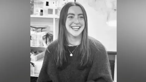 Delamar Academy A black-and-white photograph of a young woman seated indoors framed from the waist upwards. She has long, straight hair parted in the middle and is facing slightly to one side not directly towards the camera. She is wearing a dark knitted jumper and a necklace with a small pendant.