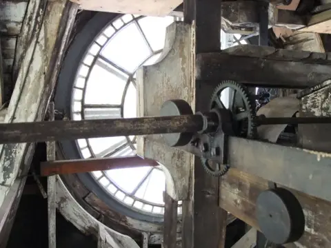South Kesteven District Council An interior view of a clock tower showing the mechanical workings, including metal gears, shafts and wooden supports. It is in front of large circular clock face with segmented glass panels.