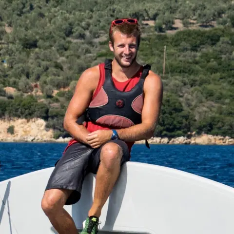 Alex Bailess A young man - Jack - is sitting on a sailing boat wearing a life jacket. 