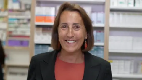 Jamie Niblock/BBC A woman smiles at the camera as she stands inside a pharmacy. She has brown hair that is cut in a bob style. She is wearing a black blazer with a red top underneath. She has dangling red earrings.