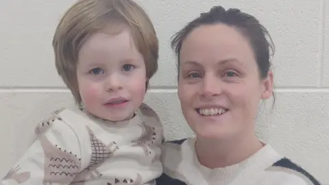 A mother hold her child in her arms - both have brown hair and are wearing similar cream and brown jumpers