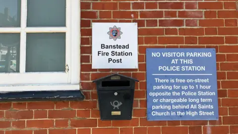 A red brick wall with a sign which says: "Banstead Fire Station Post", with a black post box below. Next to it is another sign which says: "No Visitor Parking At This Police Station. There is free on-street parking for up to 1 hour opposite the Police Station or chargeable long term parking behind All Saints Church in the High Street." There is also an edge of a window frame. 