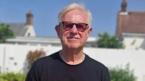 Chris Dean, who has grey hair and wears a black T-shirt, looks directly at the camera wearing sunglasses 