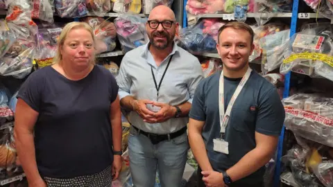 One woman and two men are standing in front of shelves full of plastic clear bags with colourful toys in them. The woman on the left has blonde shoulder length hair and is wearing a navy blue T-shirt. The man in the middle is bald with a dark moustache and beard and is wearing black-rimmed clear glasses, a lanyard and light blue shirt and jeans. The man on the right has short brown hair, is also wearing a lanyard and a navy top.