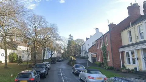 High Street in Shincliffe. It is a narrow two-lane road, with cars parked on each side. The street is lined with trees and houses on both sides.