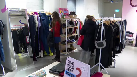 Two women browse through clothes on rails in the Thrift & Gift store next to a tall mirror