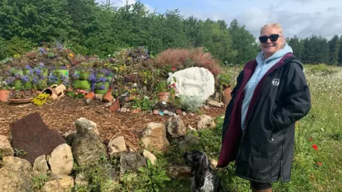 BBC/Mick Lunney A woman, Zoe Keeton, in a dark, long coat and sunglasses with a dog at her side, smiles for a photograph standing next to the mud maiden figure