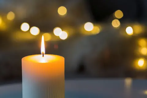 A burning cylindrical white candle in the foreground and lights behind, that are slightly blurrred.