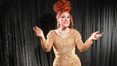 Knowsley Pride Filla wears a sequinned sparkly gold coloured dress with bejewelled necklace and ruby earrings. Filla has red hair in a beehive style and with arms slightly raised, and smiles at the camera