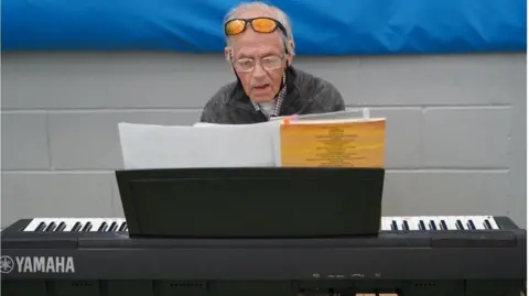 Bill Booth is sitting at an electronic piano. He is wearing glasses and has sunglasses perched on top of his head. He is reading sheet music.
