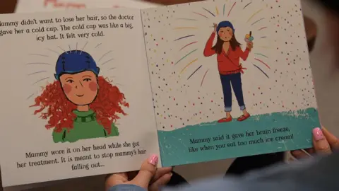 Leanne is reading the children's book. There is an illustration of a woman with curly hair wearing a cold cap. Text around it reads: "Mammy didn't want to lose her hair, so the doctor gave her a cold cap. The cold cap was like a big, icy hat. It felt very cold. Mammy wore it on her head while she got her treatment. It is meant to stop mammy's hair falling out... Mammy said it gave her brain freeze like when you eat too much ice cream!"