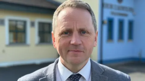 A man, with short brown hair, wearing a white shirt, navy tie and grey suit. There is a blue and yellow building behind him. 
