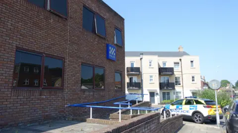 Outside of Huntingdon Police Station, it is a brick building with a sign on the side saying "POLICE" and there is a marked police car outside.