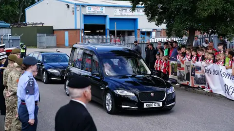 Jacob King/PA Wire A children's football team and people in military uniform line the streets while a hearse travels along the road