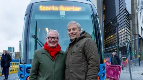 Mayor Richard Parker (left) is pictured with Jeremy Dale (right) stood in front of a blue tram. Mr Parker is wearing an army green long coat with a red wool scarf and thick black frame glasses while Mr Dale is wearing a khaki hooded waist length coat.