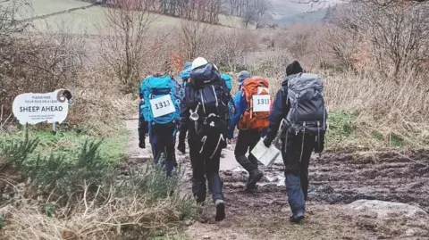Four people walking over muddy ground in the wintry Devon countryside. They all have rucksacks on and are walking together.