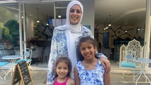 A woman with two girls standing in front of her. They stand in front of a restaurant.