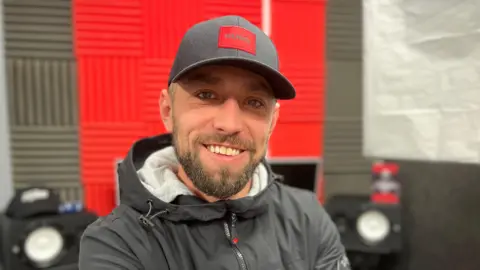 Un hombre de poco más de treinta años con barba recortada, sonriendo y mostrando los dientes. Lleva una chaqueta negra y una gorra de béisbol negra con un logo cuadrado rojo en el medio. Detrás de él se ve desenfocado el equipamiento de un estudio de grabación.