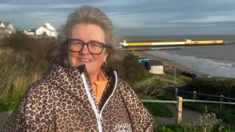 Jayn Willis wearing a leopard print jacket. Behind her is the sea and a pier. She has glasses with black frames and grey hair. She is smiling.