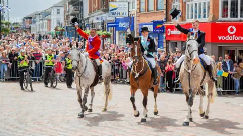 Martin McNae Photography Cornet Ross Findlater on his grey horse holding his riding hat in the air, riding alongside Cornet's Lass Robyn Peden on a bay horse holding her crop in the air and riding next to Dr Ranjit Thomas riding a grey horse holding his hat in the air. They are all riding up a cobbed street with hundreds of smiling onlookers standing against small barriers in the background